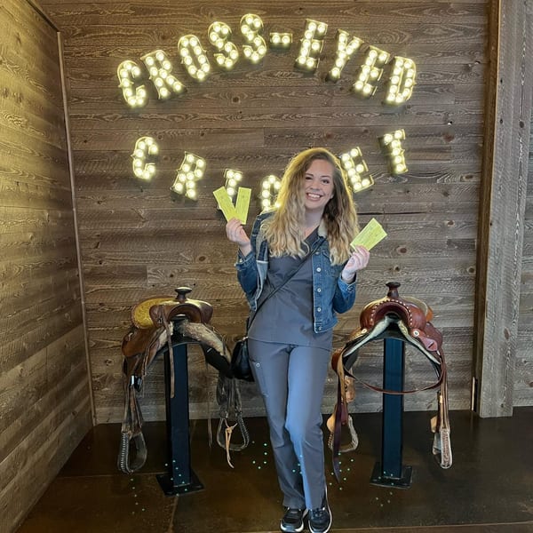 A smiling woman holds tickets in front of a lighted "Cross-Eyed Cricket" sign and two saddles.