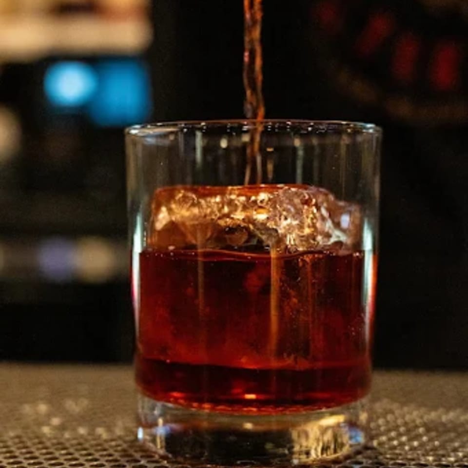 A dark amber cocktail with a large ice cube, being poured into a glass at The Cross-Eyed Cricket.
