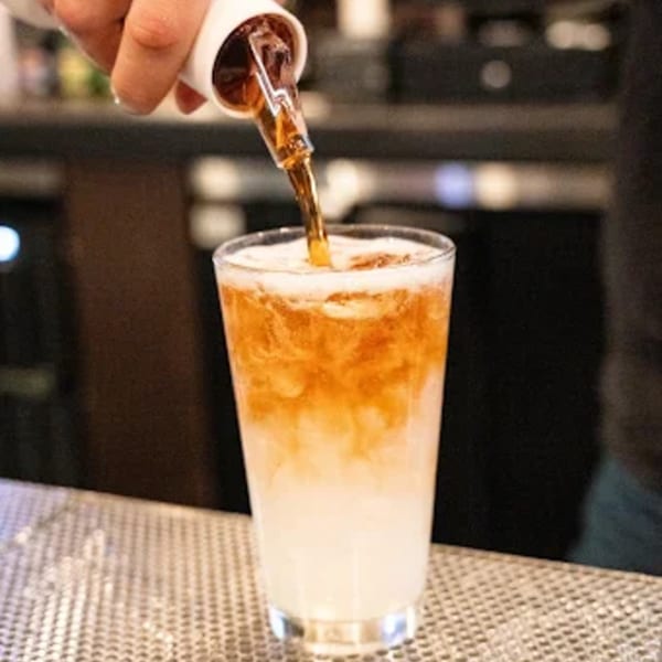 A bartender pours a dark brown liquid from a jigger into a tall glass with a white drink.