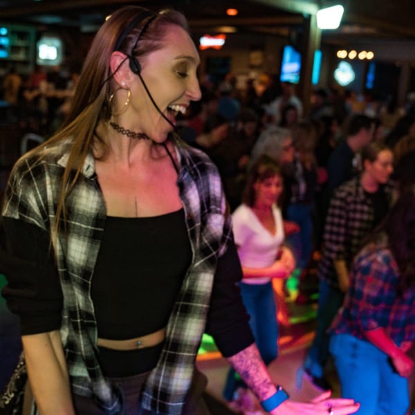 A woman with a headset sings karaoke, smiling, in a lively, dimly lit bar with other patrons.