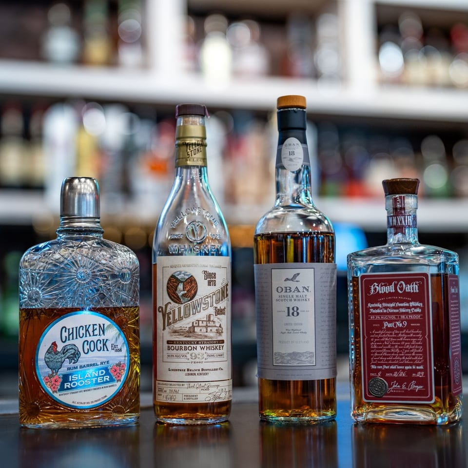 Four bottles of whiskey, including Chicken Cock, Yellowstone, Oban, and Blood Oath, on a bar.