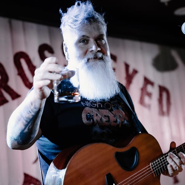 Musician with white hair and beard holding a drink and a guitar on stage at The Cross-Eyed Cricket.