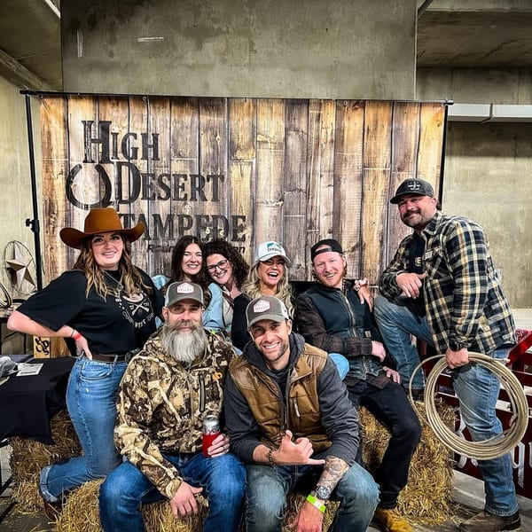 Eight people in Western wear, including cowboy hats and plaid, posing on hay bales in front of a wooden sign.