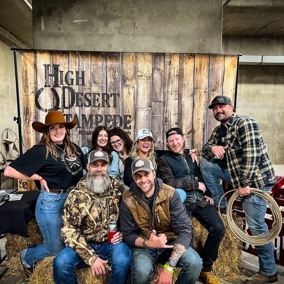 Eight people in Western wear, including cowboy hats and plaid, posing on hay bales in front of a wooden sign.