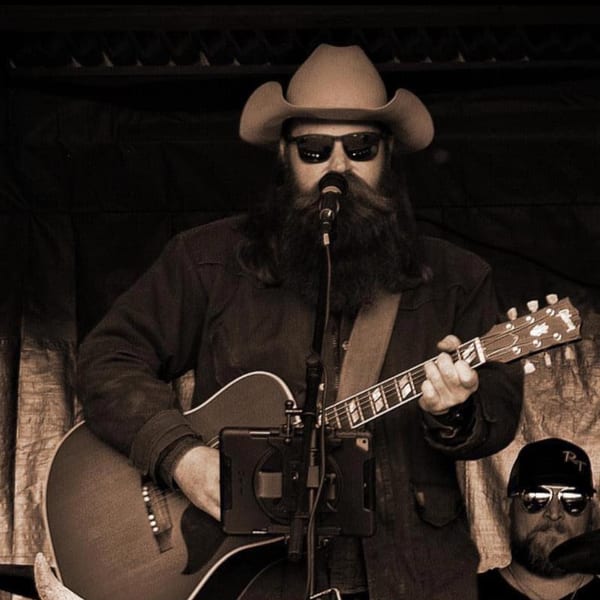 A musician with a long beard, cowboy hat, and sunglasses playing an acoustic guitar.