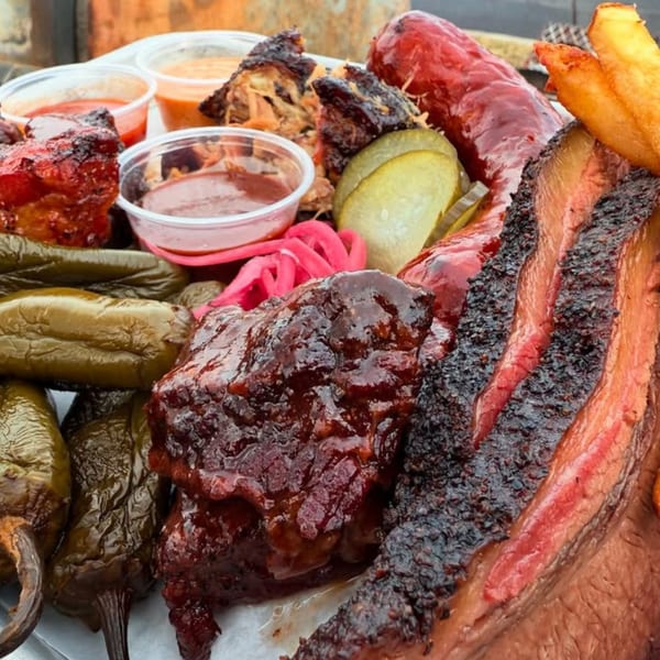 Close-up of a BBQ platter with brisket, ribs, sausage, peppers, pickles, and dipping sauces.