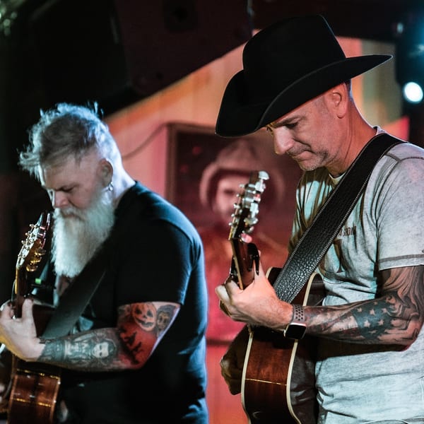 Two musicians, one in a cowboy hat, playing acoustic guitars on a dimly lit stage.