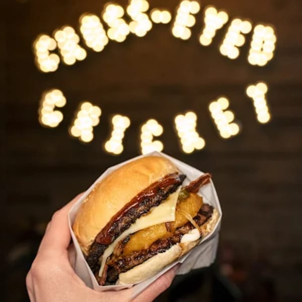 Hand holding a BBQ burger with cheese, pineapple, and bacon, with a lit-up "Cross-Eyed Cricket" sign in the background.