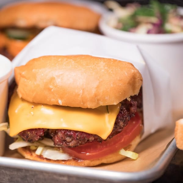 Close-up of a Cross-Eyed Cricket cheeseburger with tomato, onion, and lettuce on a metal tray.