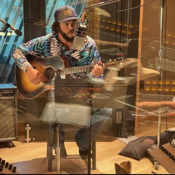 A musician in a patterned shirt and cap plays an acoustic guitar into a microphone.