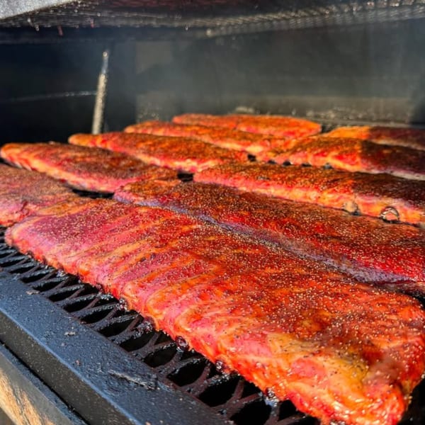 Rows of seasoned ribs cooking on a black grill inside a smoker at The Cross-Eyed Cricket.