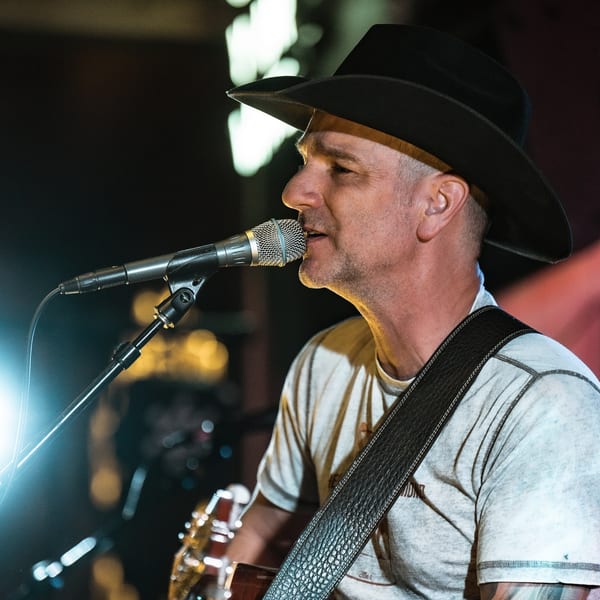 A man in a black cowboy hat sings into a microphone while playing guitar at The Cross-Eyed Cricket.